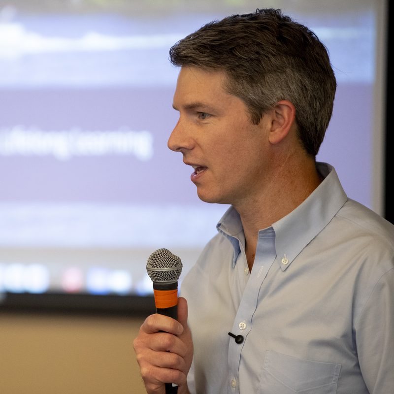 Chris Huber Reitz on stage at the Chautauqua Institution, speaking with a microphone, with an &lsquo;AI in Society and Lifelong Learning&rsquo; slide visible behind him