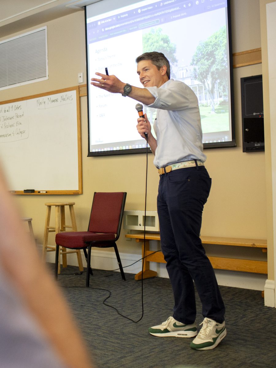 Chris Huber Reitz speaking at the Chautauqua Institution, full-body shot mid-gesture with microphone in hand, agenda slide visible on the screen behind him